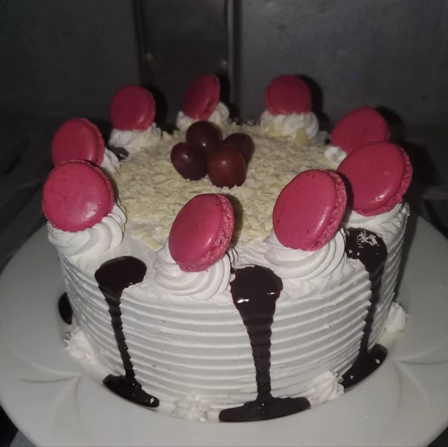 White cake topped with pink macarons, white chocolate shavings and chocolate drip